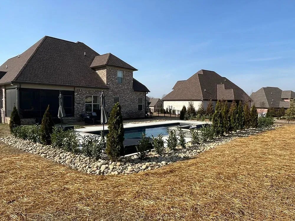 Backyard with a pool, landscaping, and a large brick house under a clear blue sky.
