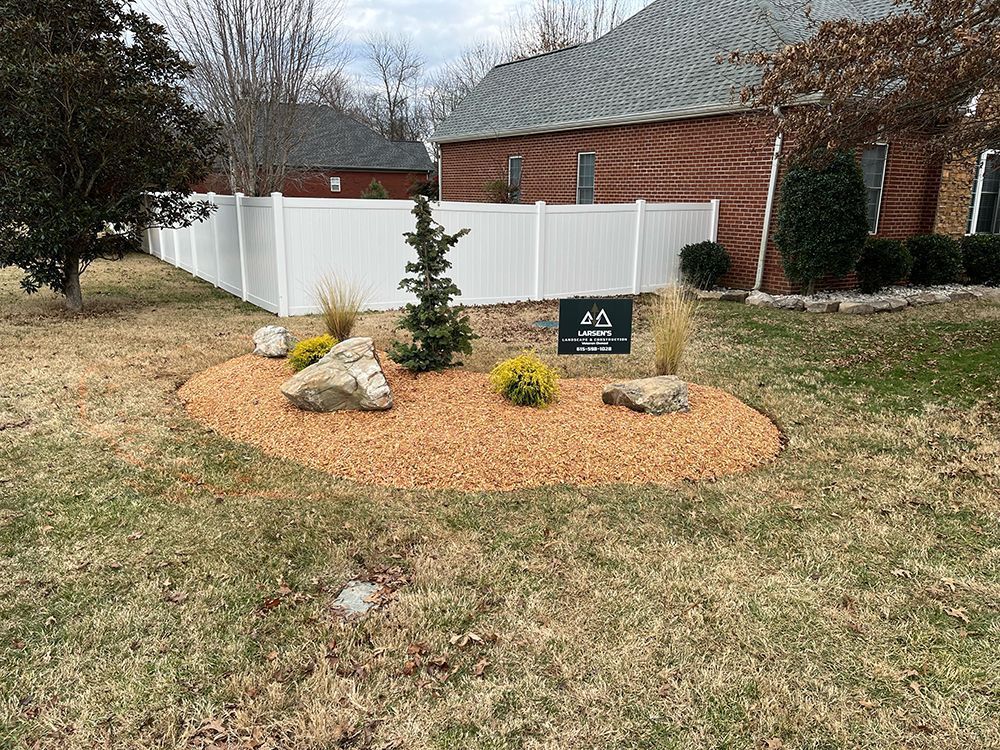 A white picket fence surrounds a landscaped yard with a sign, rocks, mulch, and shrubs in front of a brick house.