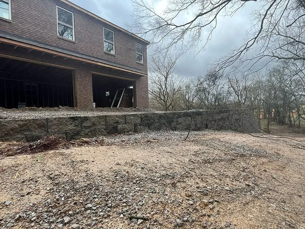 Two-story brick house under construction, overlooking a gravel yard and retaining wall, overcast sky.