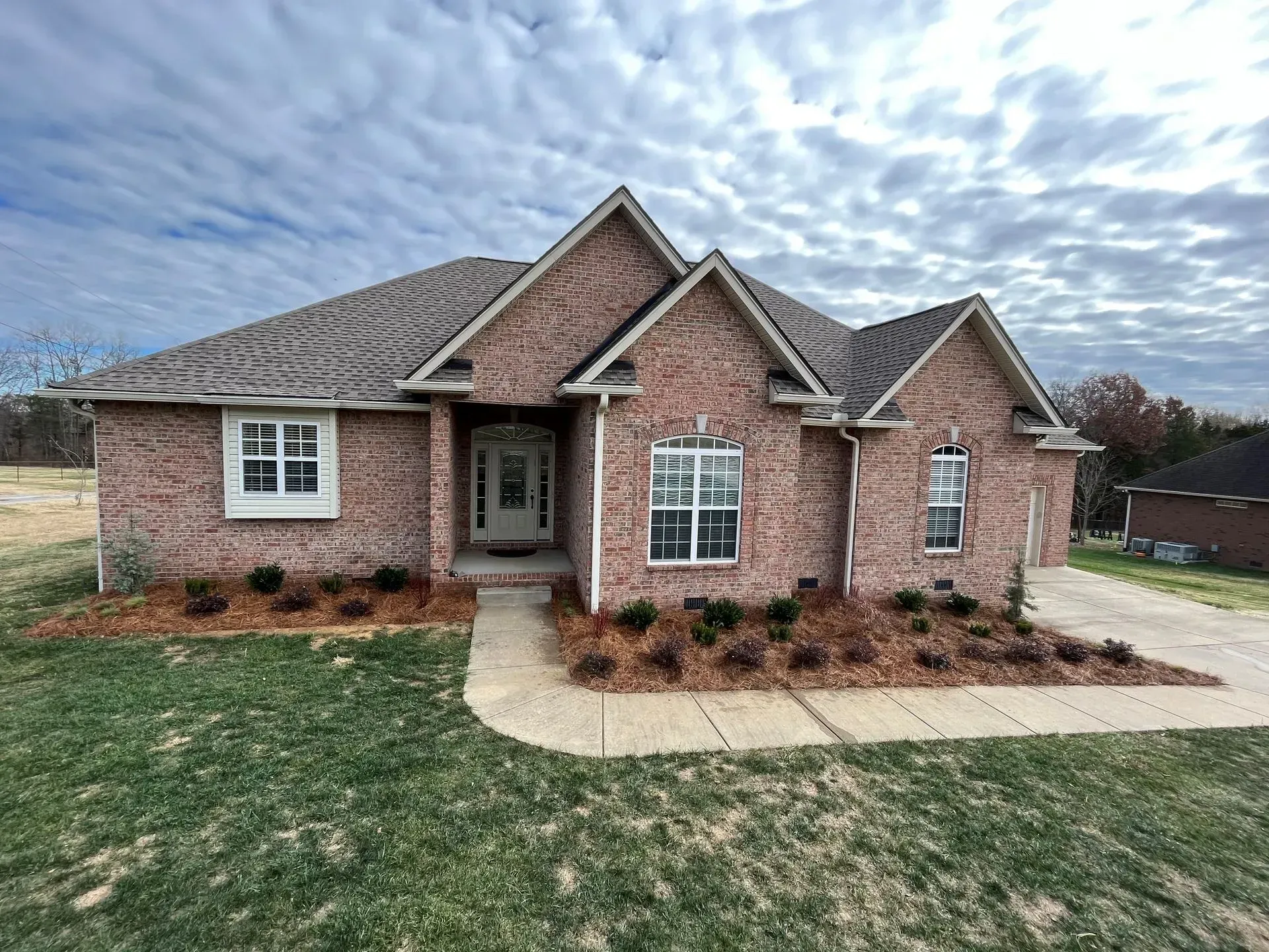 Brick house with a gray roof on a cloudy day. Green grass and a concrete walkway in front.