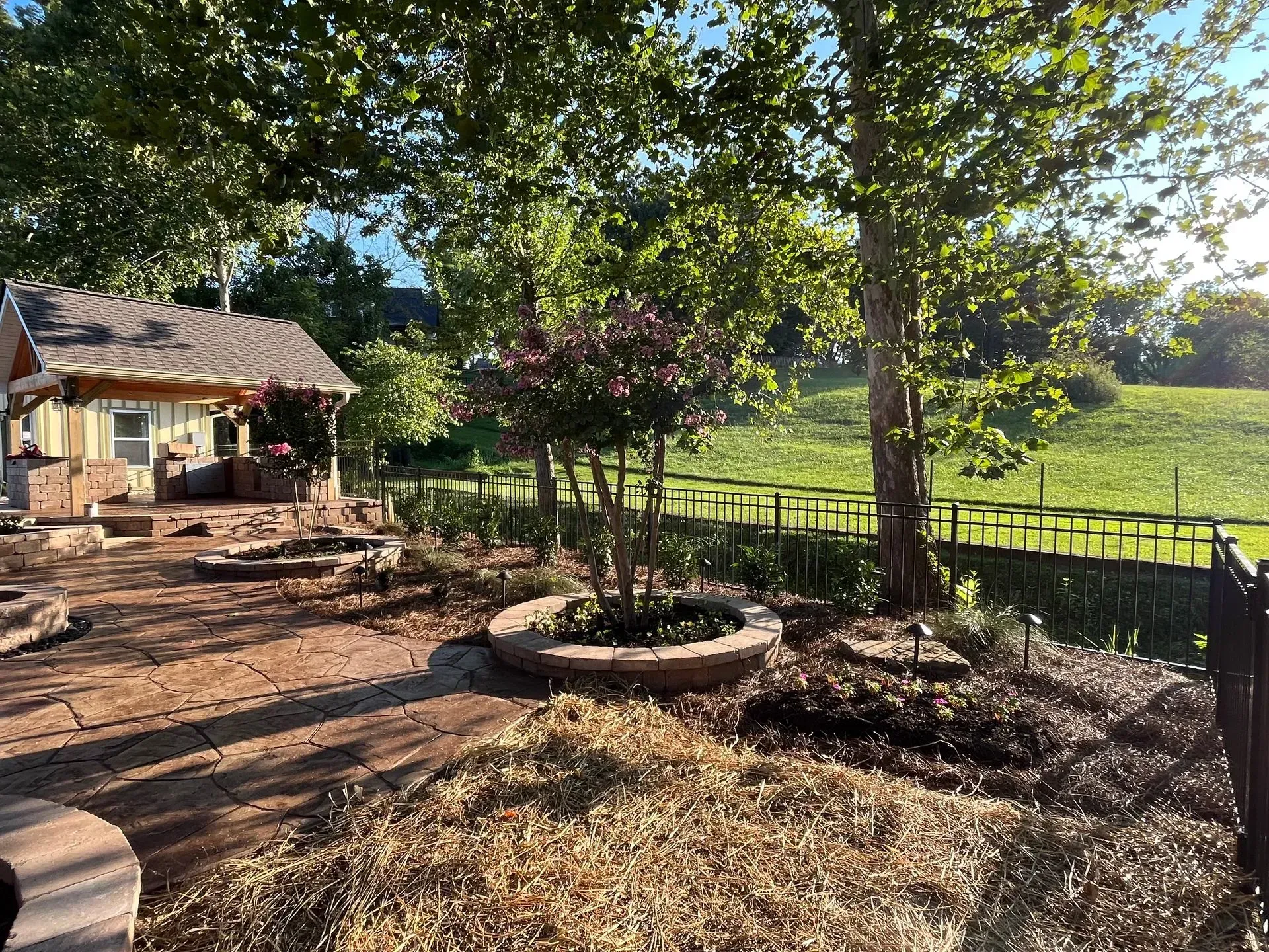 A backyard scene with a small house, brick patio, trees, a flower bed, and a grassy hill.