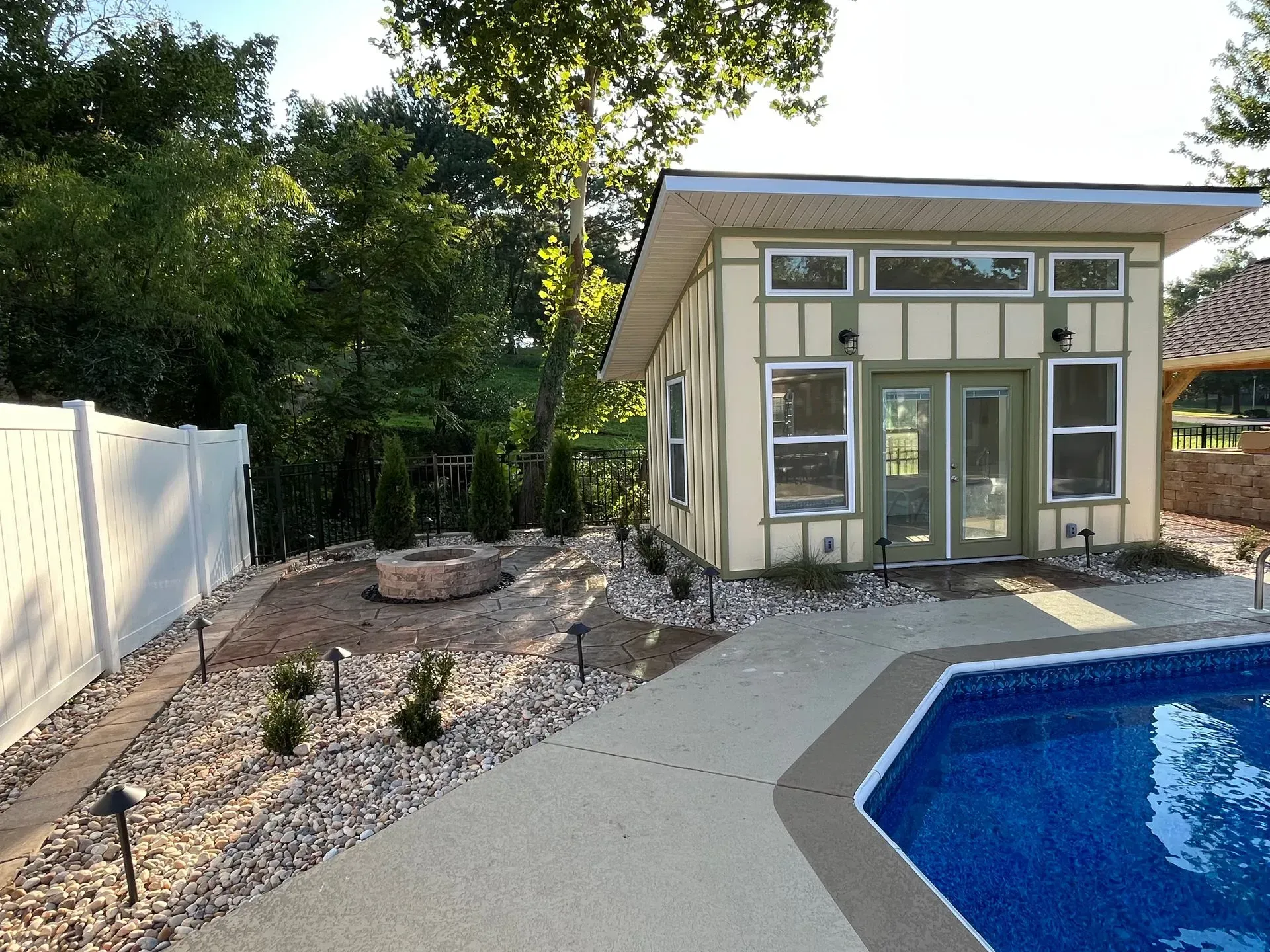 Poolside cottage with light green trim, a stone path, fire pit, and white fence.