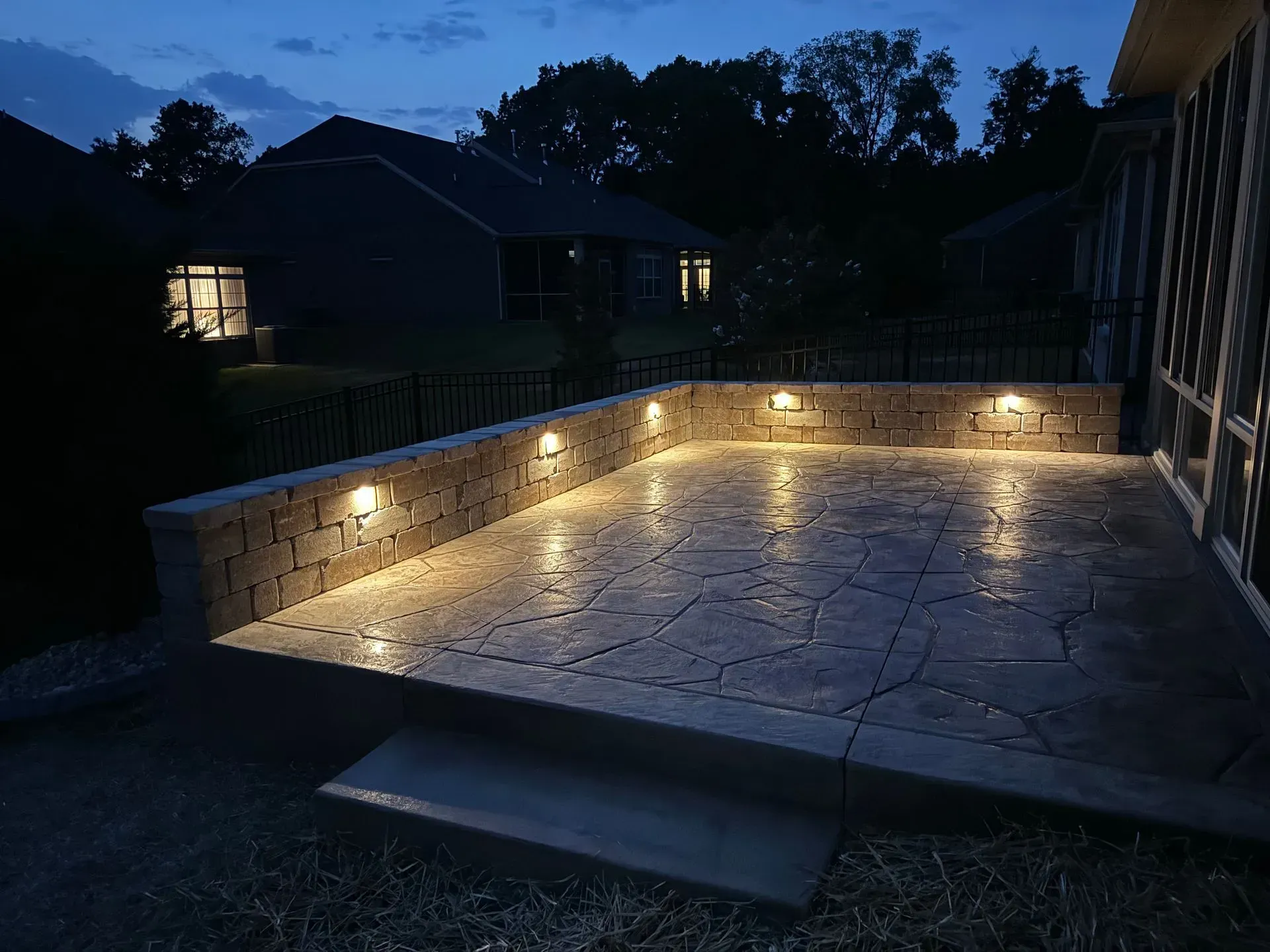 A patio with built-in lights at night. Brick wall, dark gray floor. Houses in the background.