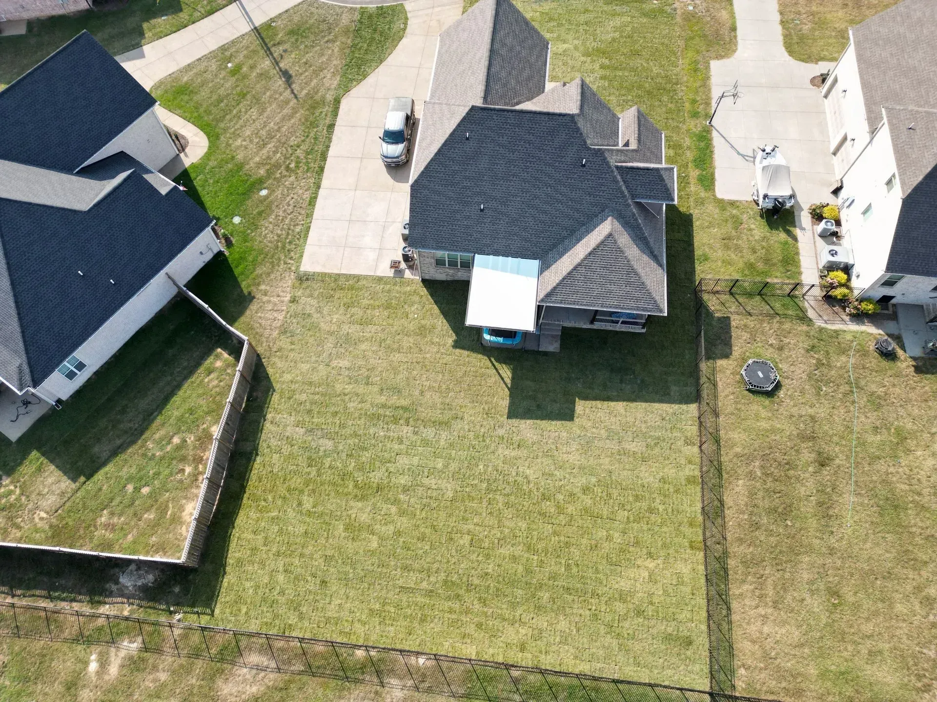 Overhead view of houses with gray and black roofs, green lawns, and a fence.