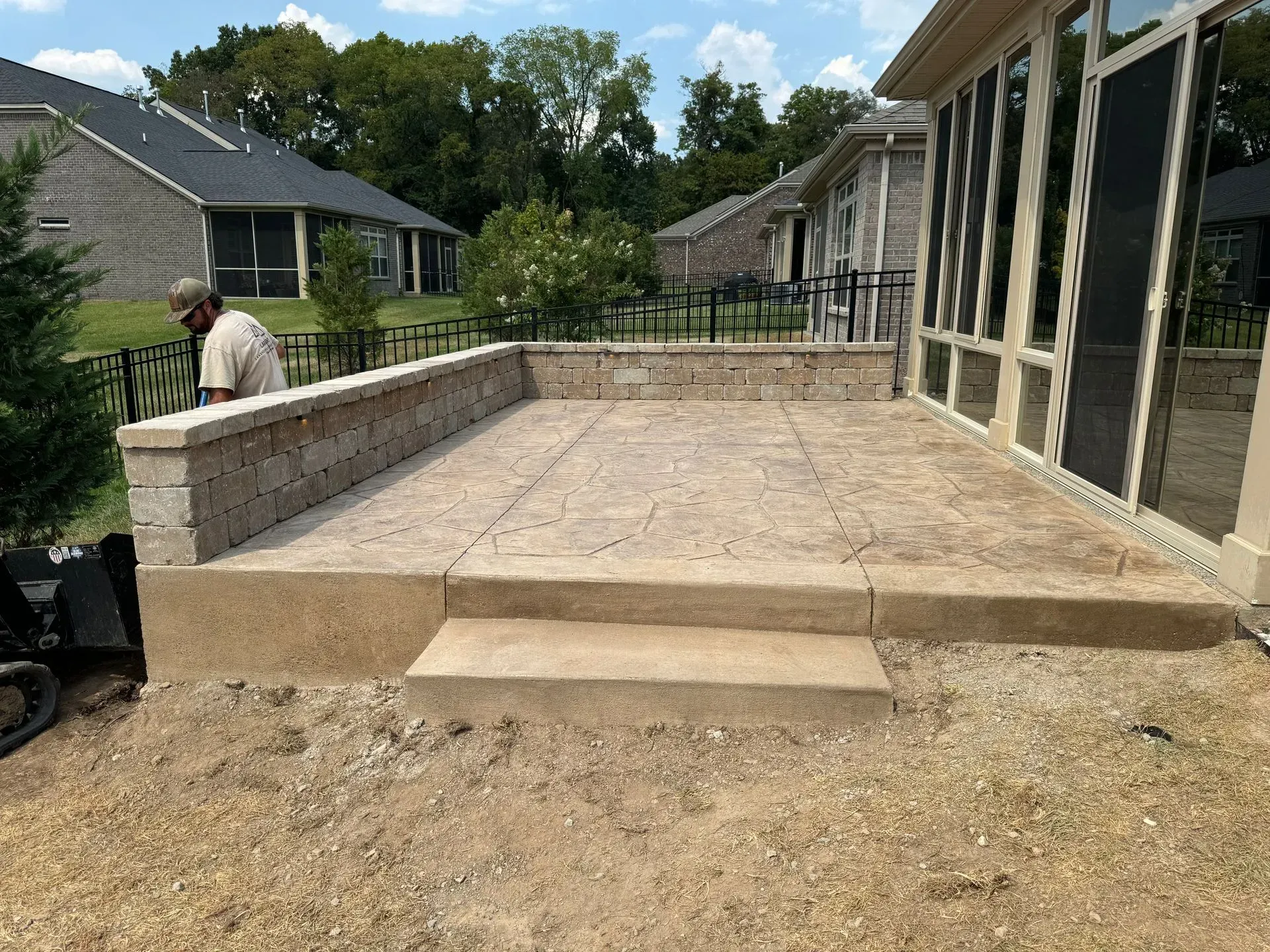 A man works on a concrete patio with steps and retaining wall next to a sunroom.