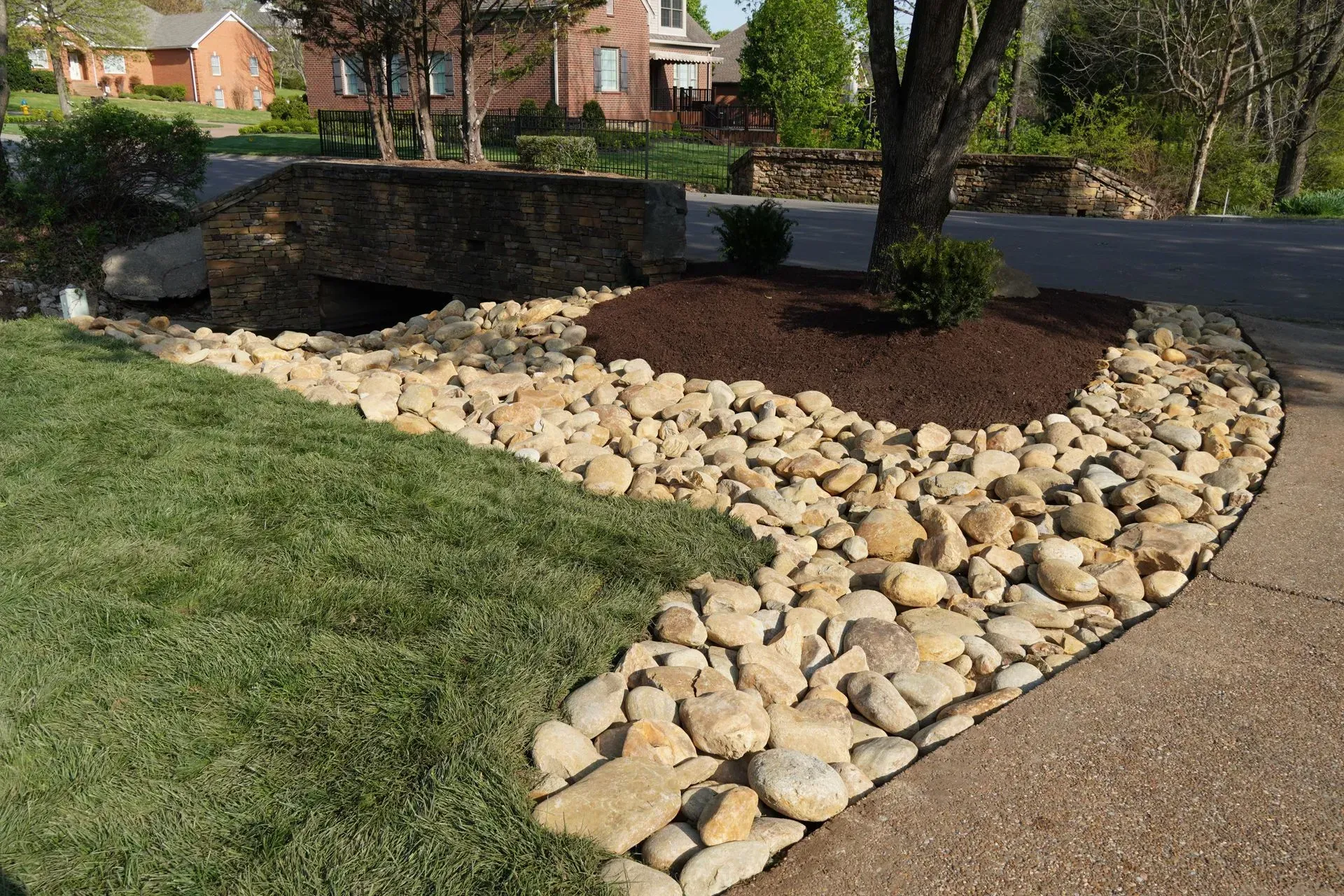 A landscaped yard with grass, rocks, and a tree next to a road and sidewalk.