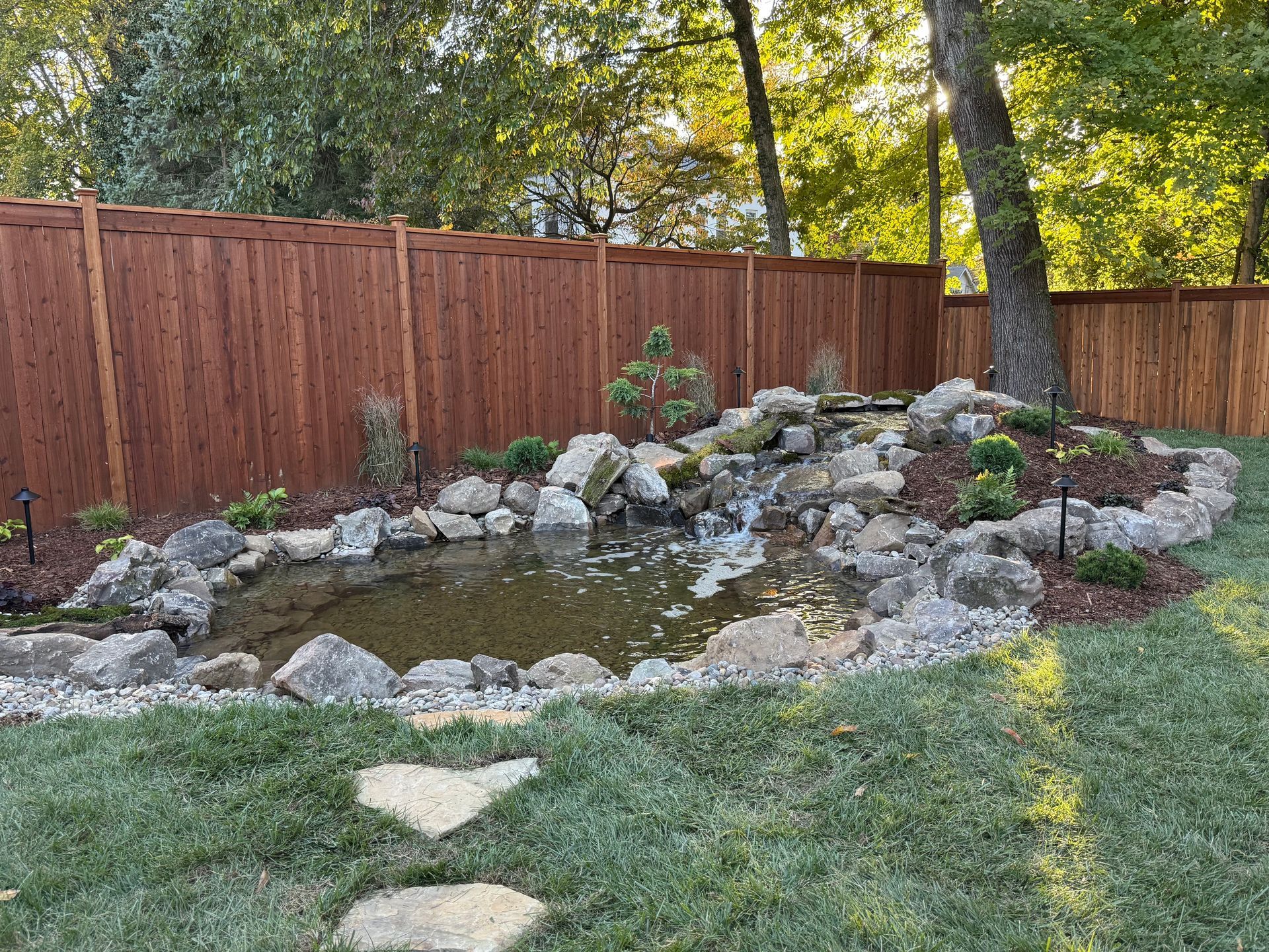 Backyard pond with waterfall, surrounded by rocks and plants, wooden fence in background.
