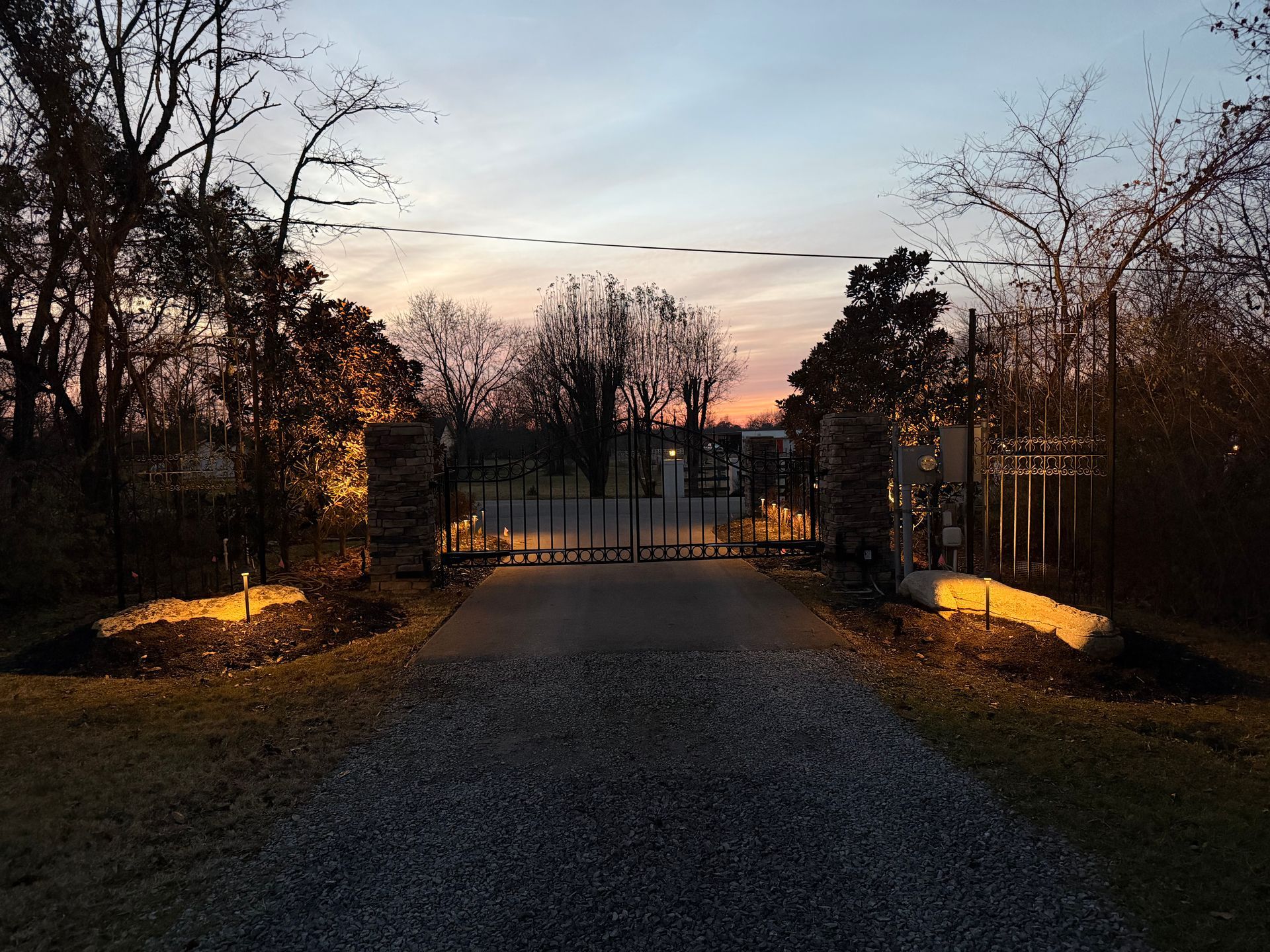 Gated entrance to a road at dusk; stone pillars, illuminated by yellow lights, flank black iron gates. Trees in the background.