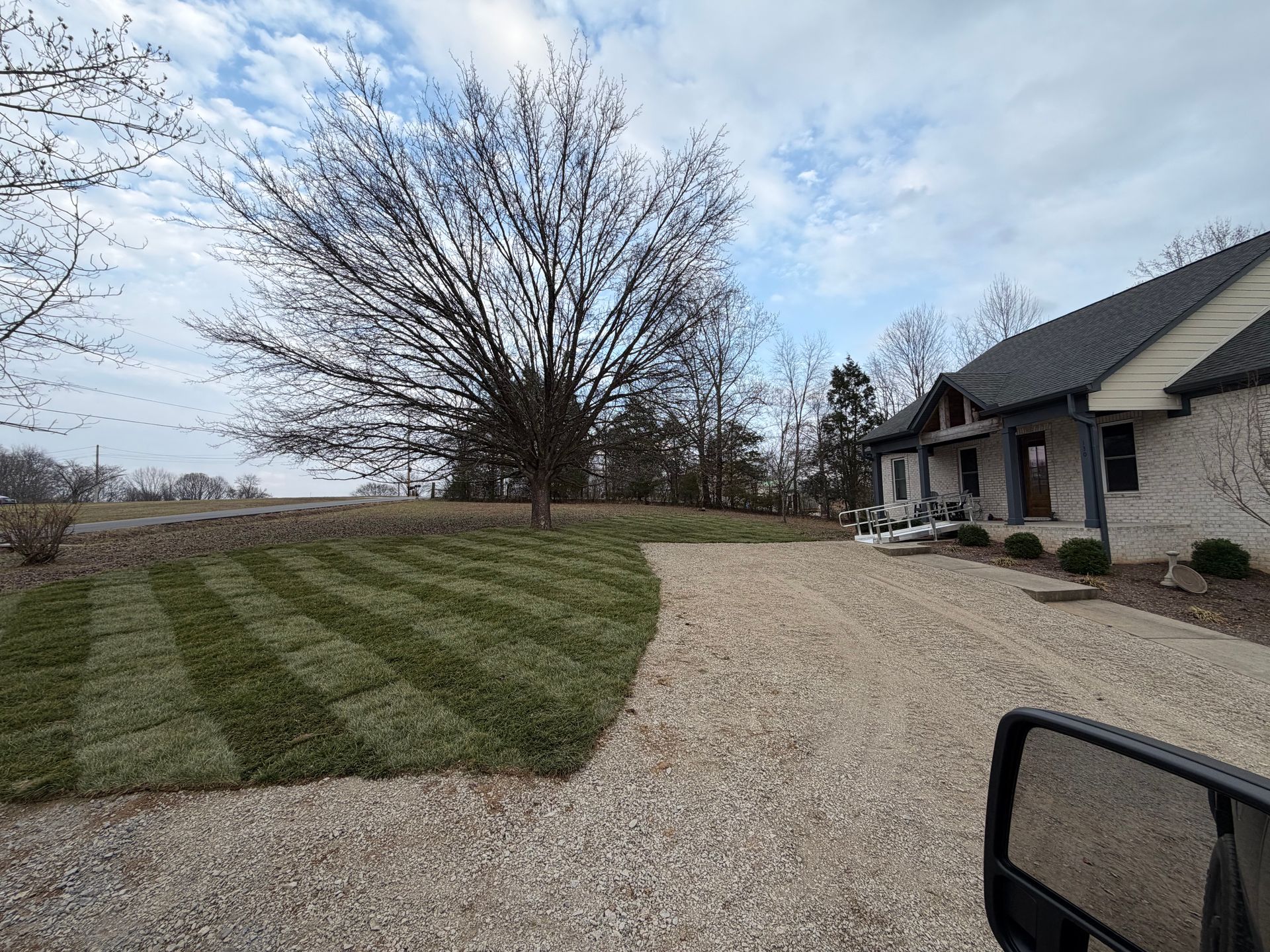 Lawn with striped pattern, gravel driveway leading to a house, bare tree in the background, cloudy sky.