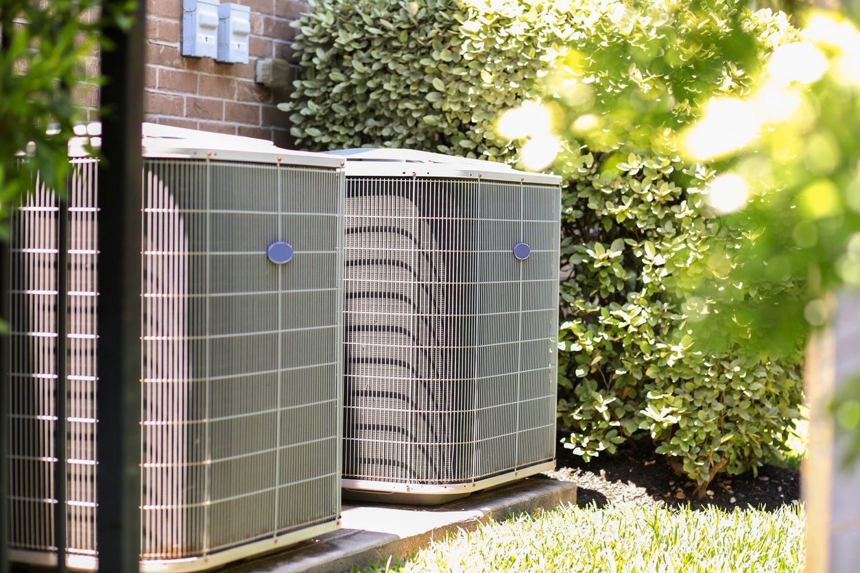 Two air conditioning units next to a brick wall, surrounded by greenery.