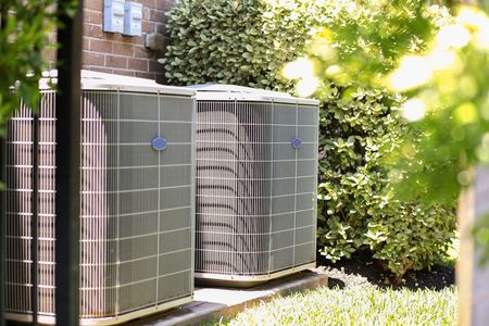 Two outdoor air conditioning units against a brick wall, surrounded by green bushes.