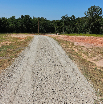 A gravel driveway leads through a cleared, grassy lot toward a dense treeline under a clear blue sky.
