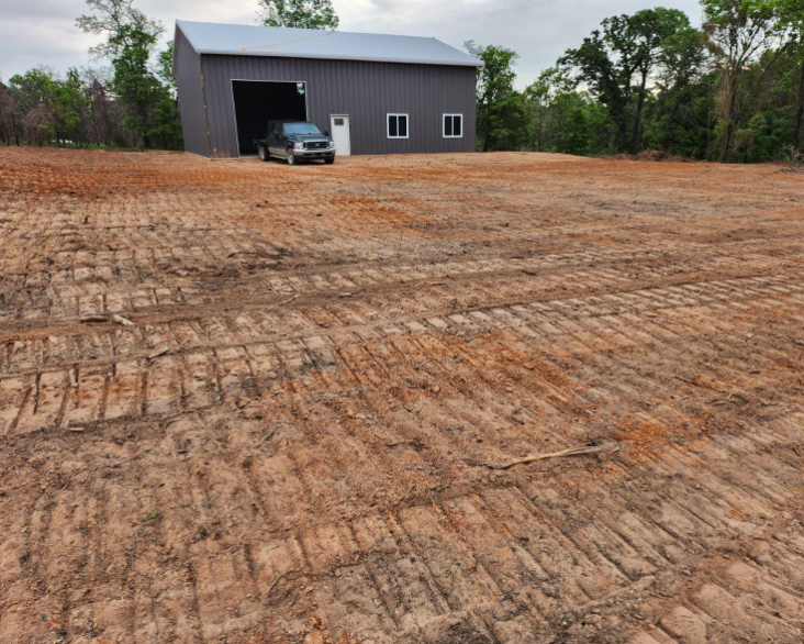 A dark metal barn stands on a freshly cleared dirt lot with prominent tire tracks and surrounding trees.