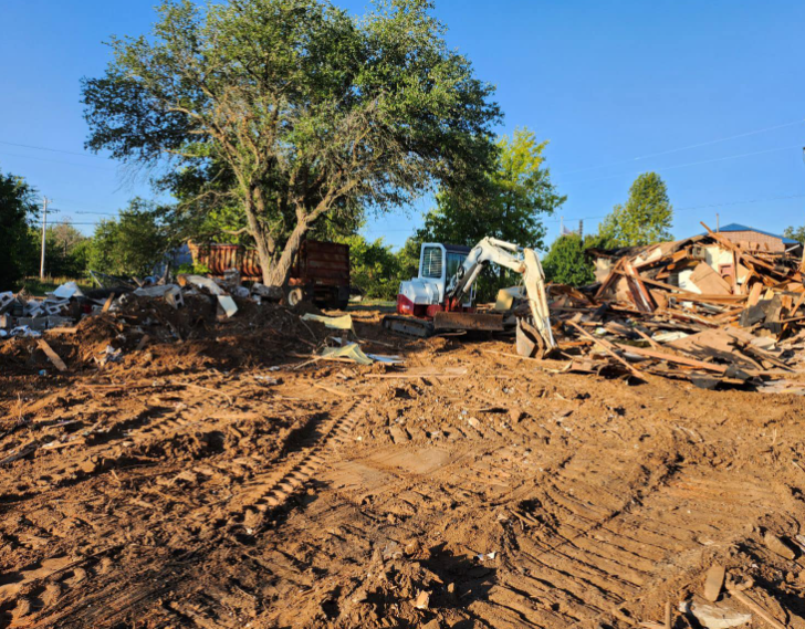 A white excavator sits on a muddy, uneven construction site amidst piles of debris and a large tree under a clear blue sky.