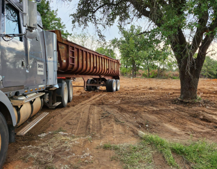 A grey semi-truck with a large, rust-colored dump trailer parked on a dirt lot near a tree under an overcast sky.
