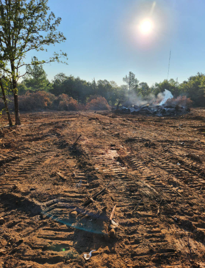A sunlit clearing shows churned, muddy ground from heavy machinery with a pile of burning debris and smoke in the distance.