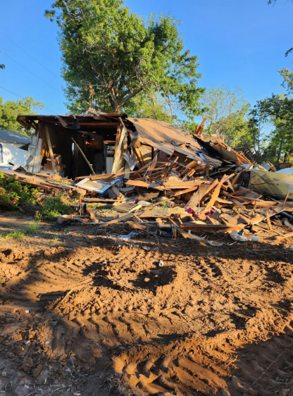 A demolished wooden building sits under a clear blue sky, with heavy vehicle tracks visible in the dirt in the foreground.