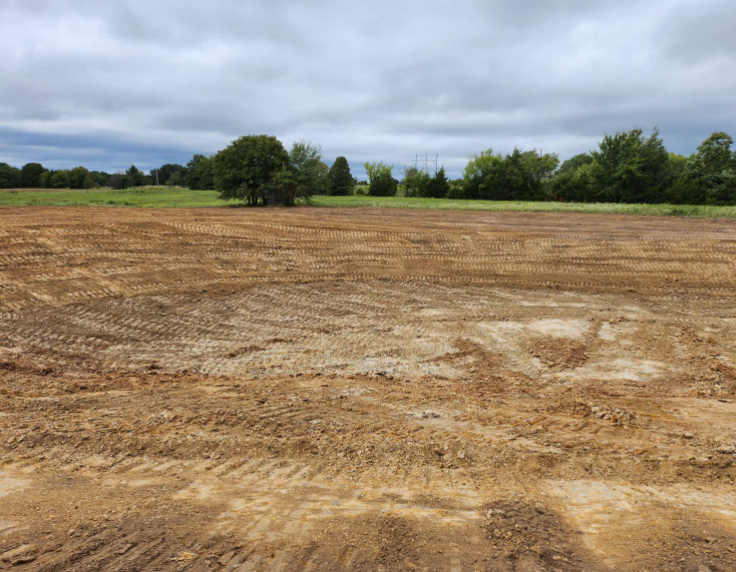 A wide, graded dirt lot with tire tracks in the foreground and a line of green trees under a cloudy sky.