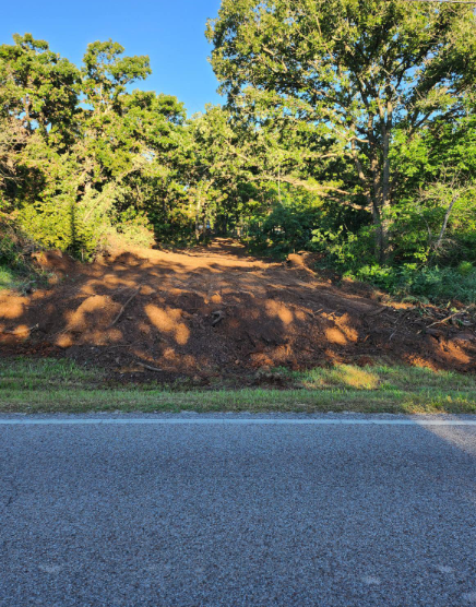 A section of land with freshly cleared red soil, positioned behind a paved road and bordered by a forest of green trees.