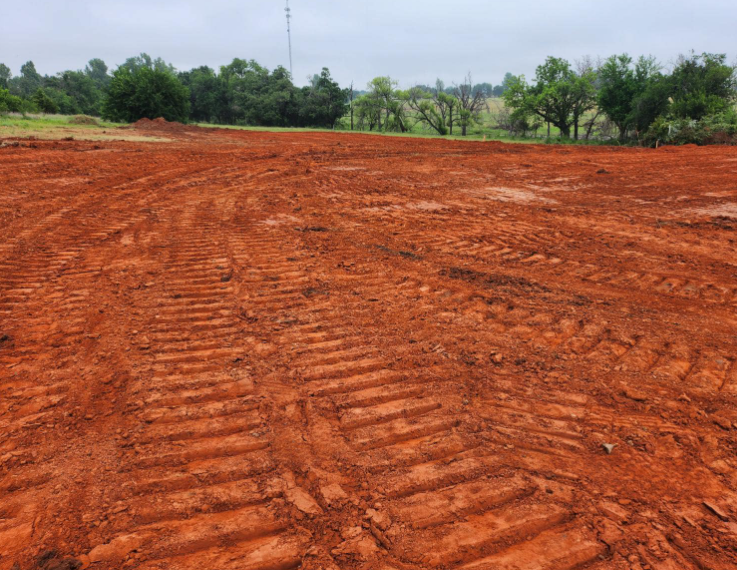 A clearing of red dirt showing heavy machinery track marks, with trees and a cloudy sky in the background.