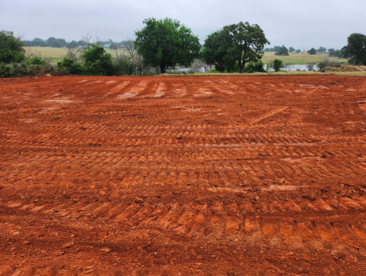 Freshly graded red dirt field with heavy machinery tire tracks, featuring trees and a pond in the background.