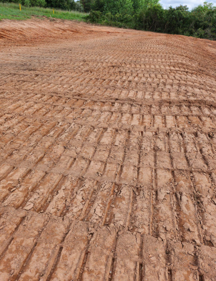 A view of freshly graded reddish-brown soil with prominent, parallel tread marks from heavy construction machinery.