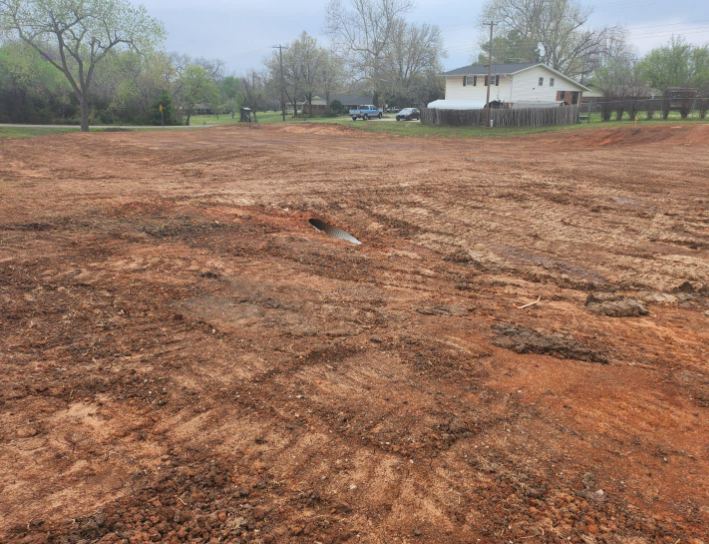 A cleared, uneven dirt lot with tire tracks in the foreground and a house visible in the background.