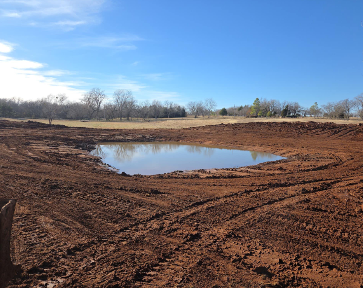 A small, muddy pond sits in the center of an excavated, red-dirt construction site under a clear blue sky.