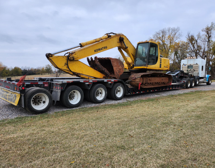 A large yellow excavator sits on a flatbed trailer being hauled by a white semi-truck in a grassy outdoor area.