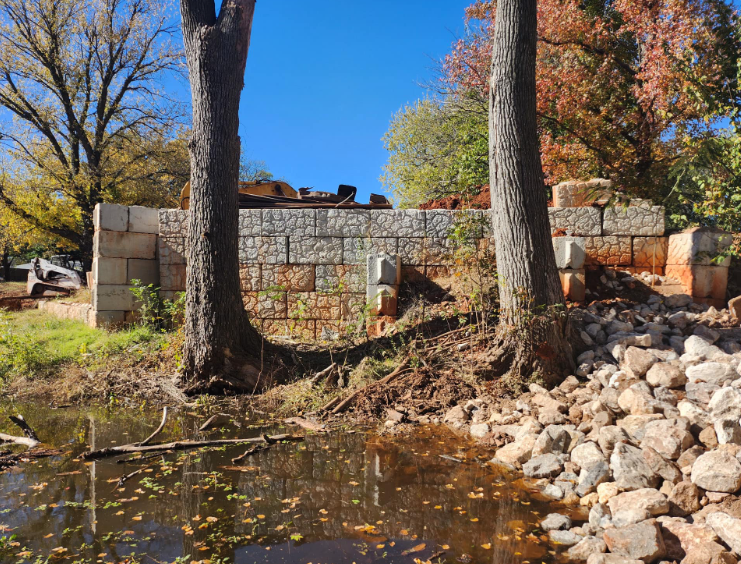 A stone retaining wall stands between two trees above a rocky shoreline at the edge of a pond.