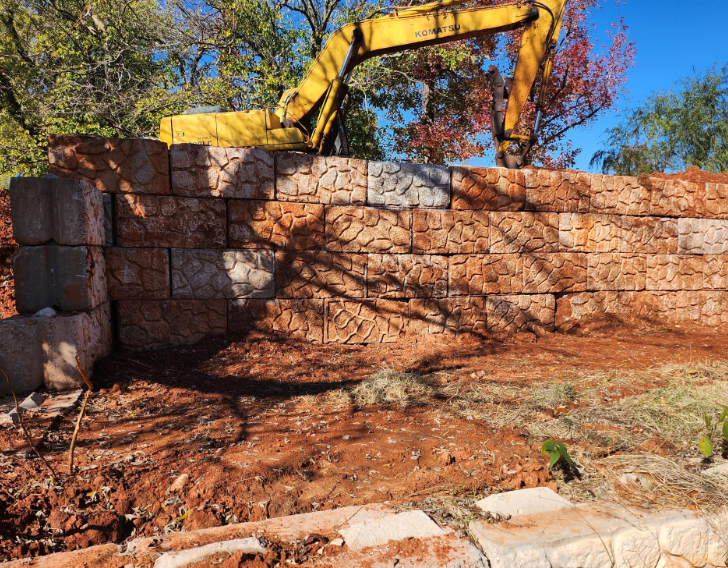 An excavator building a retaining wall with large, textured, reddish-brown stone blocks under a clear blue sky.