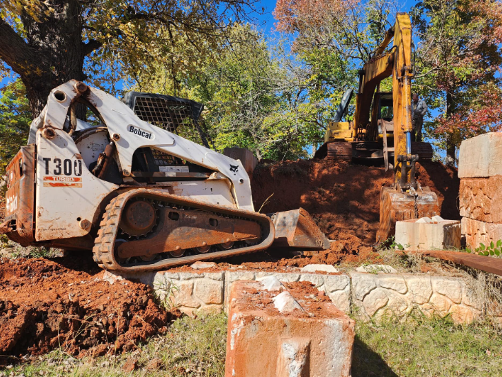 A white Bobcat T300 skid steer and a yellow excavator work on a dirt mound at a construction site with trees nearby.