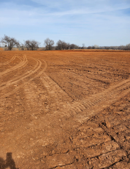 Freshly tilled red-brown soil in a vast open field with tire tracks and a distant treeline under a bright blue sky.