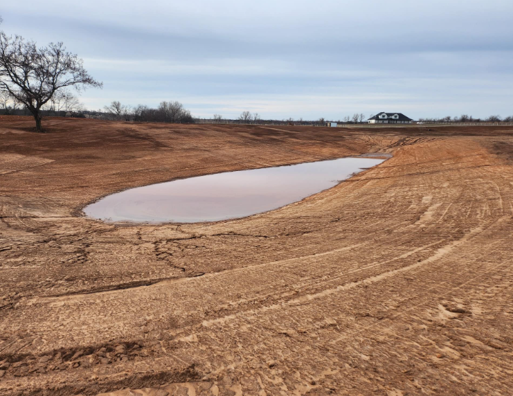 A muddy, man-made pond sits in the center of an excavated, brown landscape under a cloudy, overcast sky.