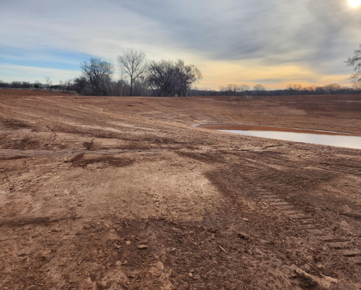 A wide, barren field of red-brown dirt with tire tracks and a small patch of water under a cloudy sunset sky.