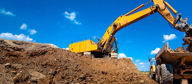 A yellow excavator loads dirt into a dump truck against a bright blue sky.