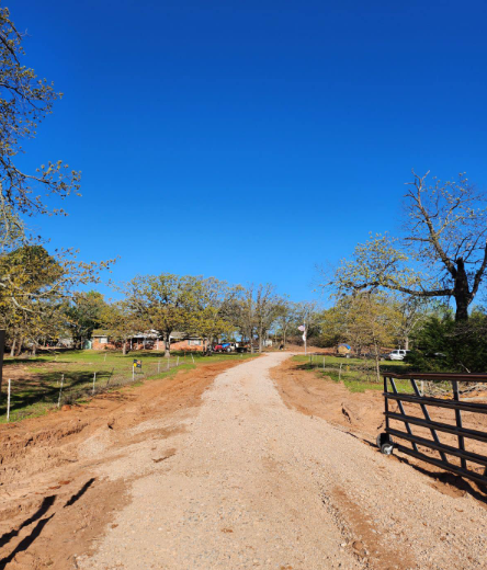 A gravel driveway leads through a rural landscape with trees, green grass, and a wooden fence under a clear blue sky.