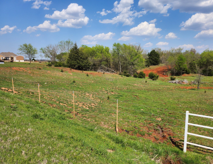 A sloping, grassy field with patches of red clay soil, thin wooden fence posts, and a white gate under a blue, cloudy sky.