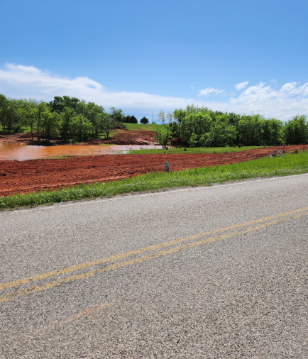 A paved road leads past a patch of exposed red clay soil and a murky, flooded area, bordered by lush green trees.