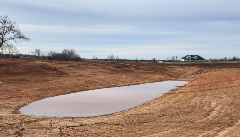 A muddy, excavated landscape under an overcast sky featuring a small pool of standing water and a house in the distance.
