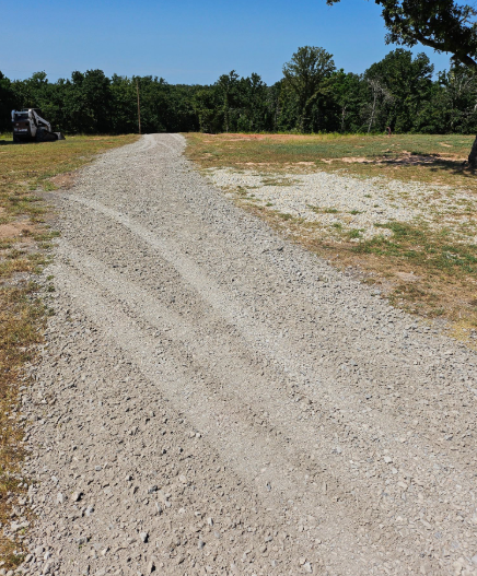 A gravel driveway curves through a dry, grassy field toward a line of trees under a clear blue sky.