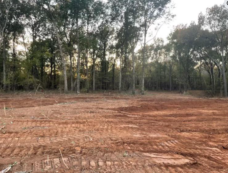 A cleared, dirt lot with visible heavy equipment tire tracks, bordered by a dense line of trees in the background.