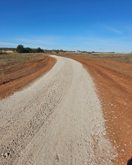 A wide, curved gravel road leads through a dry, open field with reddish-brown soil under a clear blue sky.