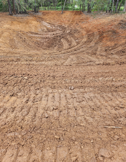 Freshly excavated reddish-brown dirt forming a basin, marked by deep tire tracks, with trees in the background.