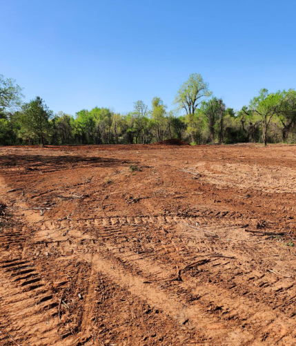 A cleared, red-dirt field with heavy equipment tracks in the foreground, bordered by a line of trees under a blue sky.