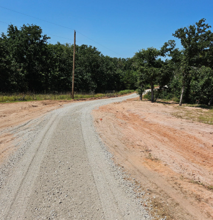 A winding gravel road stretches through a sunny landscape bordered by trees and dirt ground under a clear blue sky.