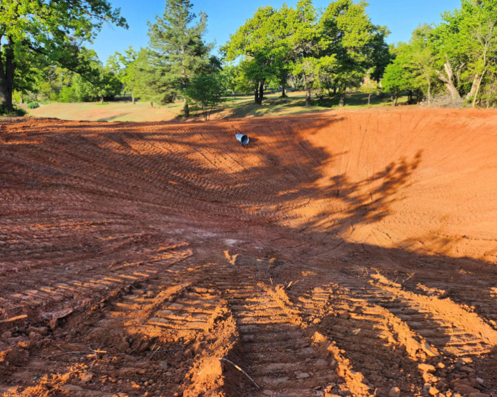 A newly excavated pond basin with red clay soil, tire tracks in the foreground, and green trees in the background.