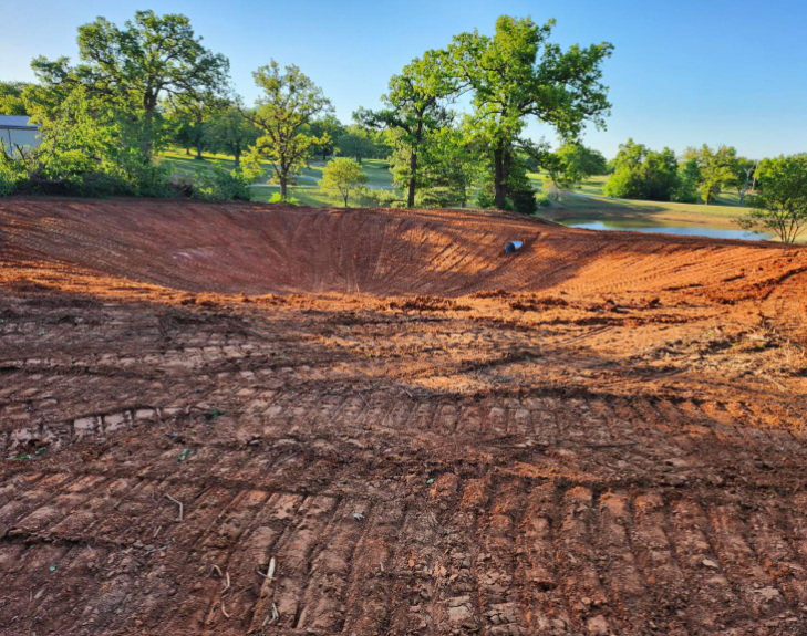 Freshly excavated, red clay earthen pond under a clear blue sky with surrounding trees and a nearby body of water.