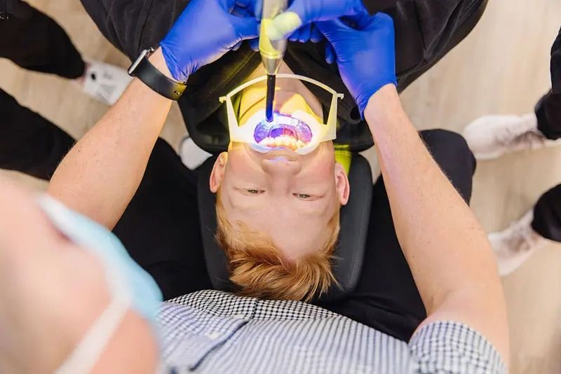 Dentist using a curing light on a patient's mouth. Patient in chair, blue gloves visible.