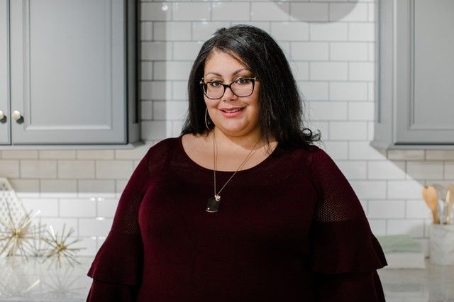 Tiffani wearing glasses and a maroon shirt is standing in a kitchen .